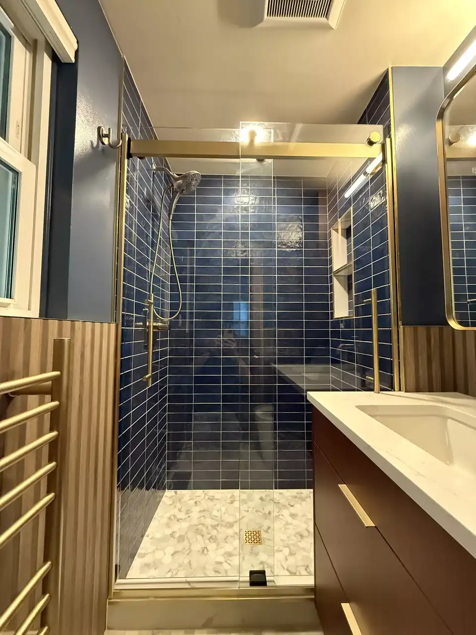 Bathroom remodeling Redmond WA by Remont: navy subway tile walk-in shower with brass-framed glass door, pebble floor, wood slat wall and heated towel rail