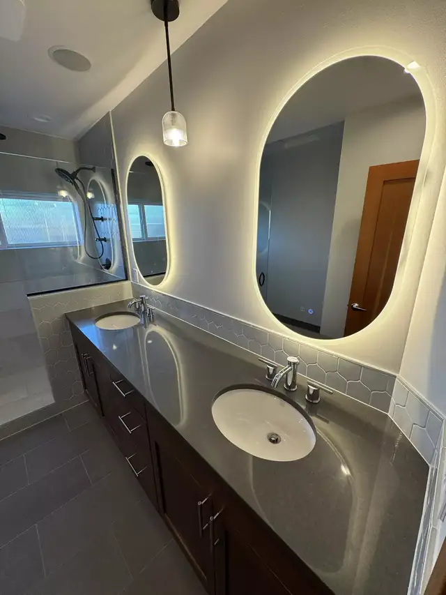 Bathroom renovation with a long dark wood double vanity, gray quartz top and backlit oval mirrors