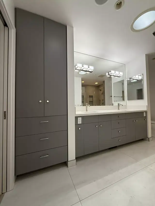 Bathroom with a tall gray linen cabinet, gray double vanity, large mirror and skylight