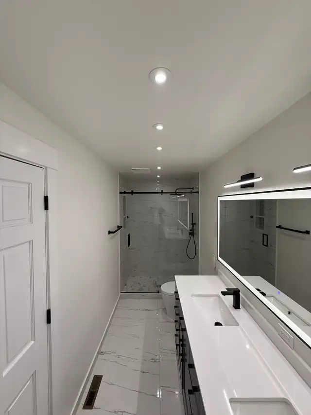 Long bathroom with white double vanity, lighted mirror and a sliding glass shower at the far end
