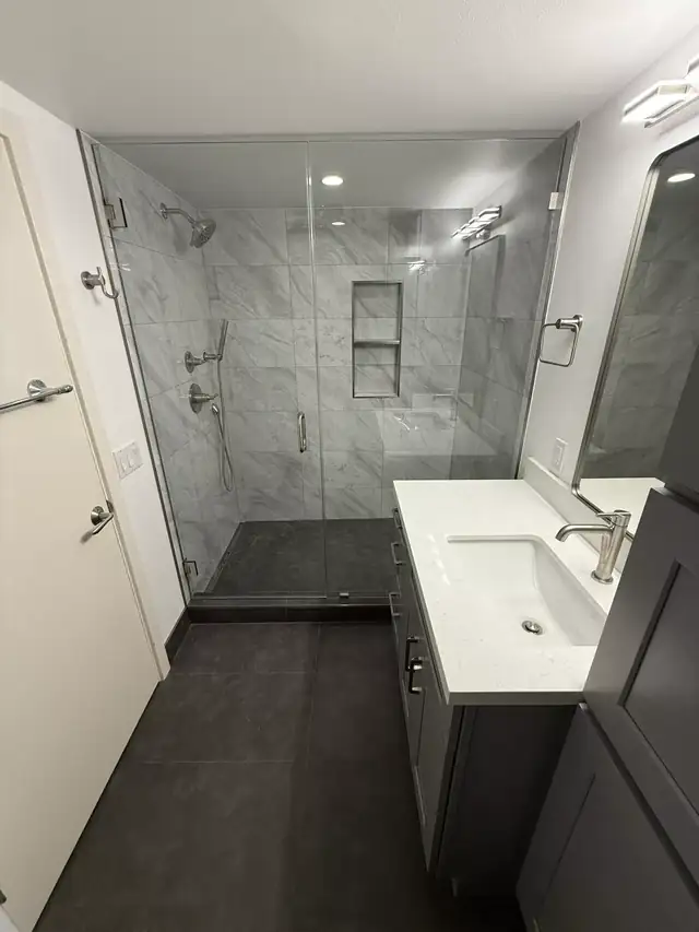 Glass walk-in shower with marble-look tile beside a white vanity and dark porcelain floor