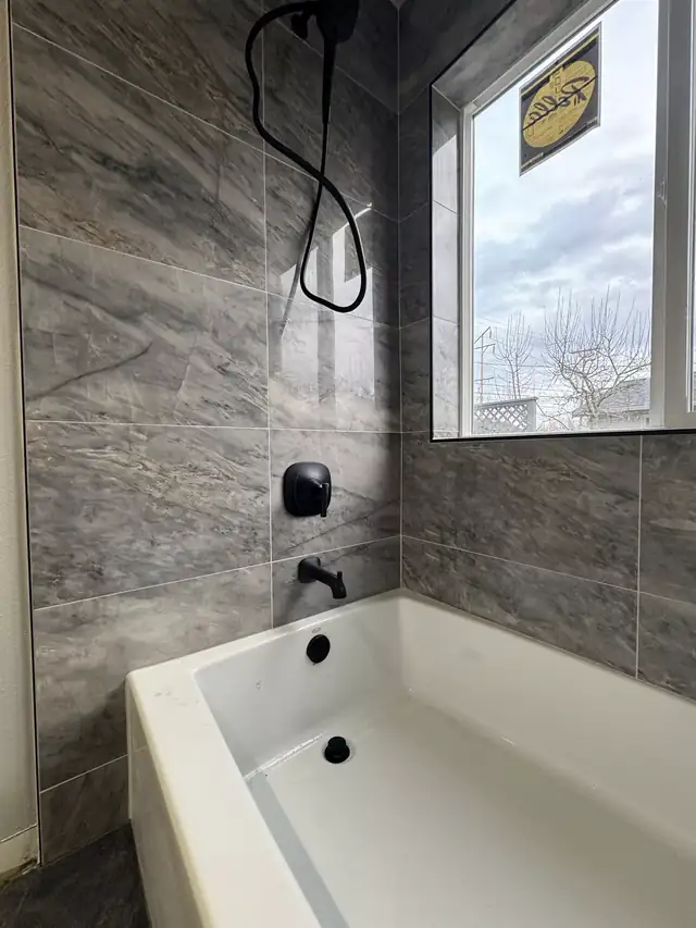 Soaking tub under a window with dark gray marble-look wall tile and matte black fixtures